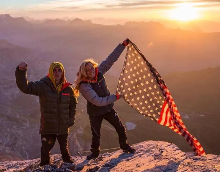 Două femei stau pe o stâncă la apus, una ridicând un braț și cealaltă ținând un steag american întins.