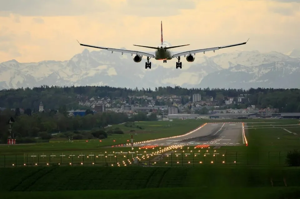 Un avion de pasager zboară pe cer deasupra unei piste de aterizare cu lumini de ghidare aprinse, având munți și un oraș în fundal.
