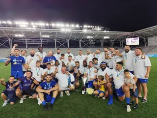 Un grup de sportivi în echipamente albastre și albe poartă medalii aurii și posează un trofeu pe un teren de sport sub reflectoare.