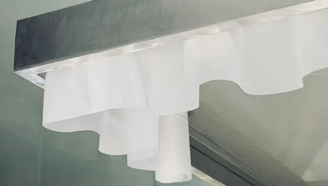 O sculptură albă din material textil sau subțire este suspendată sub o structură gri de formă dreptunghiulară.