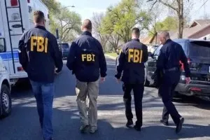 Patru bărbați purtând jachete cu inscripția FBI merg pe un drum lângă mașini și un vehicul alb mare.