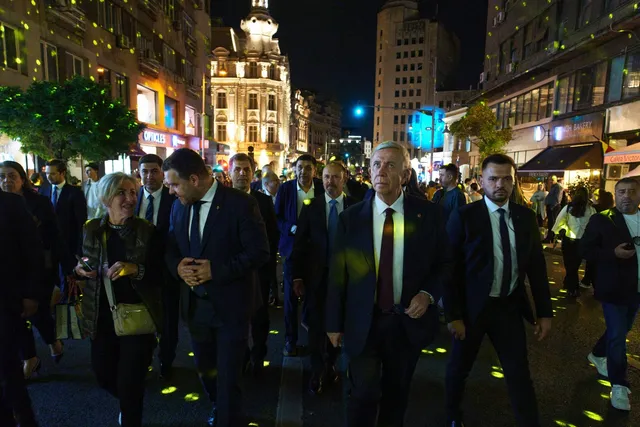 Un grup de bărbați în costume închise merge pe un trotuar iluminat de puncte de lumină verzi sub clădiri urbane noaptea.