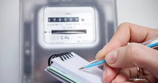 O persoană ține un stilou albastru deasupra unui mic bloc de note albe și verzi lângă un contor electric digital.
