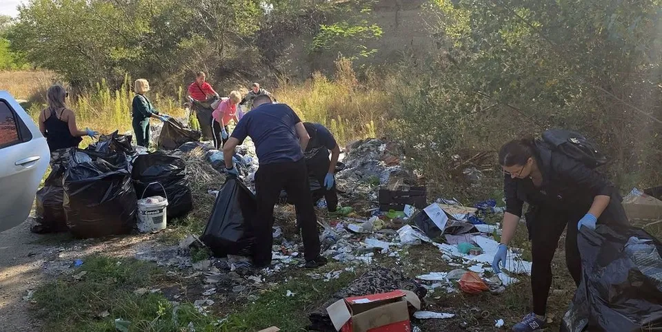 Oameni adună deșeuri în saci negri lângă o mașină, pe un drum mărginit de vegetație.