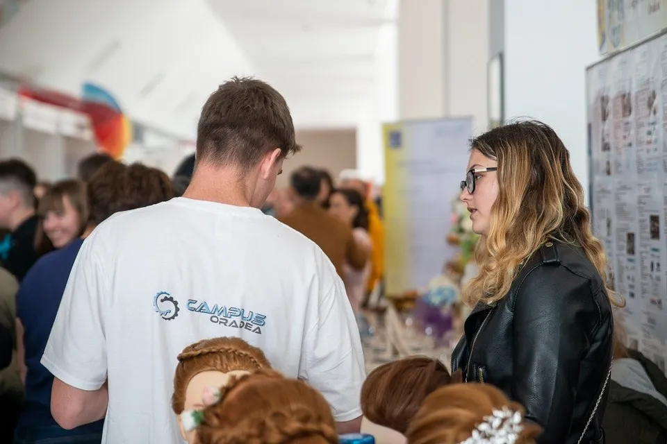 O femeie cu ochelari și o geacă neagră privește spre un bărbat cu un tricou alb pe care scrie CAMPUS ORADEA într-un spațiu interior.