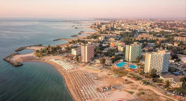 O vedere aeriană a unei zone de coastă cu clădiri, plaje cu umbrele și apă de culoare albastră sub un cer senin.