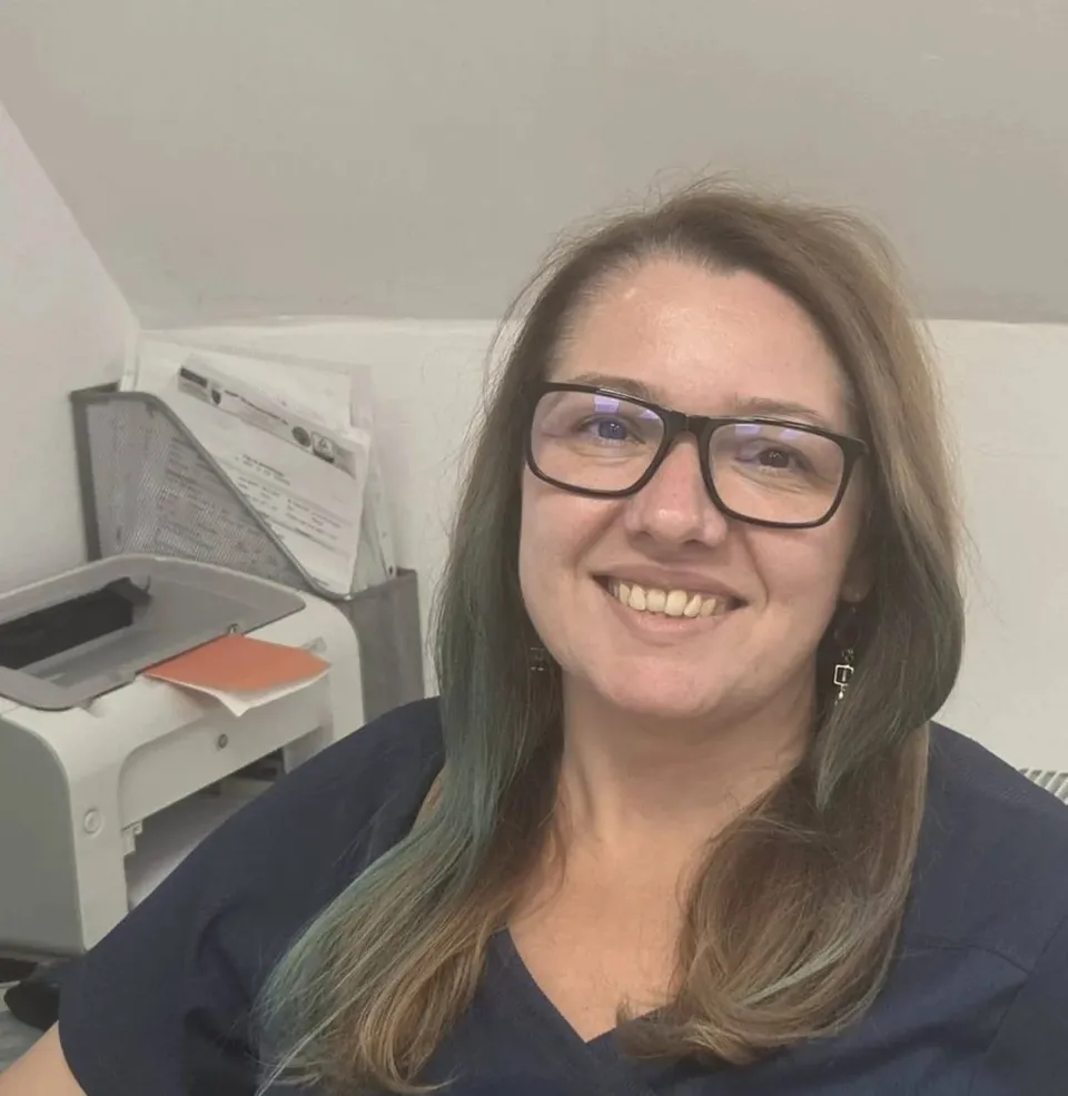 O femeie cu ochelari și părul lung, purtând un tricou albastru, zâmbește în fața unui aparat de imprimantă și a unor hârtii pe un birou.