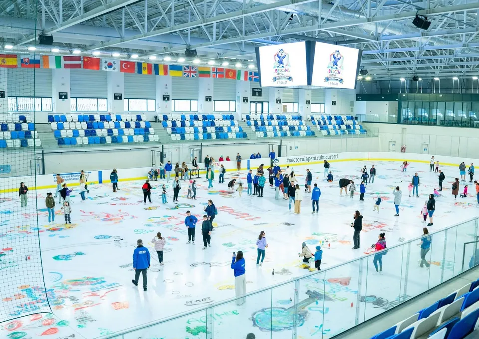 Oamenii patinează pe o pistă de gheață interioară sub un plafon metalic, în fața unor tribune cu scaune albastre și albe.