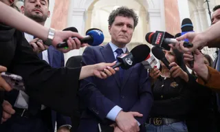 Un bărbat într-un costum albastru este înconjurat de microfoane și mâini care țin dispozitive de înregistrare în fața unui fundal de clădire.