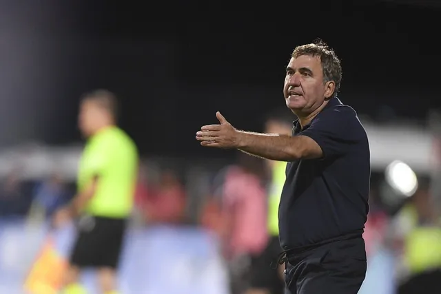 Un bărbat cu tricou închis la culoare stă în picioare și întinde mâna dreaptă într-o poziție de comandă pe un teren de sport.
