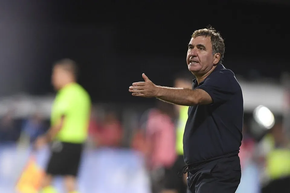 Un bărbat cu tricou închis la culoare stă în picioare și întinde mâna dreaptă într-o poziție de comandă pe un teren de sport.