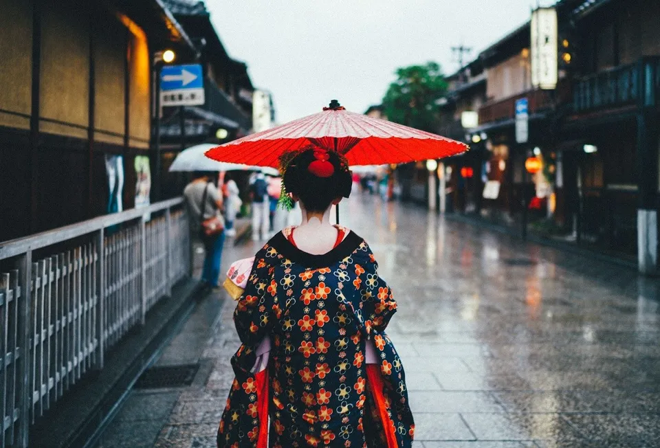 O persoană cu părul negru poartă un kimono decorat și ține o umbrelă roșie pe un trotuar umed lângă clădiri tradiționale.