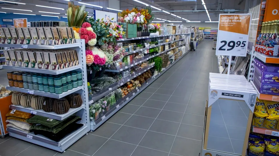 Rânduri de rafturi albe conțin flori colorate, plante și diverse produse de îngrijire, dispuse de-a lungul unui culoar de magazin cu podea gri.
