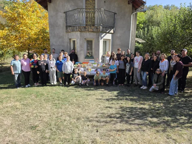 Un grup de oameni stă poziționat în linie pe iarbă în fața unei clădiri, lângă o masă cu diverse obiecte colorate.