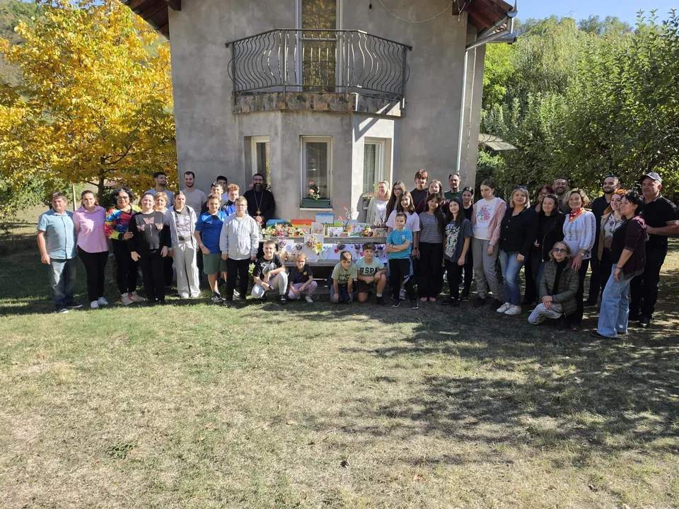 Un grup de oameni stă poziționat în linie pe iarbă în fața unei clădiri, lângă o masă cu diverse obiecte colorate.