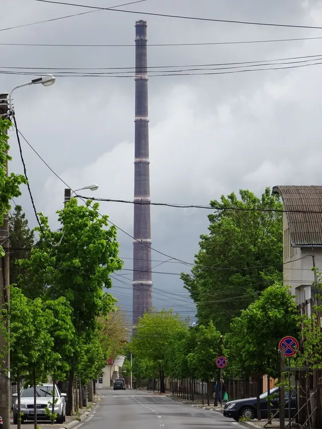 Un turn înalt și subțire se ridică deasupra unor copaci verzi, de pe o stradă cu mașini parcate și linii electrice suspendate.
