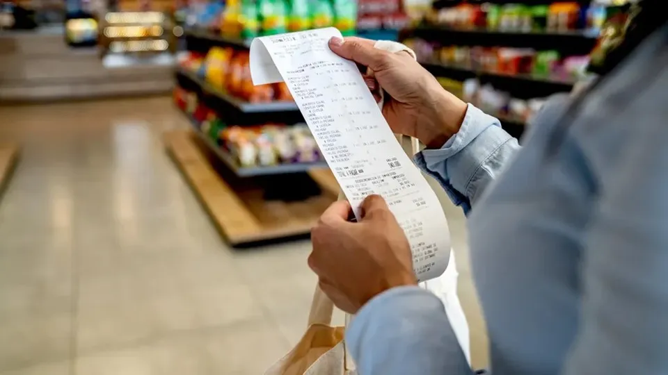 O persoană ține un bon de plată alb într-un magazin cu rafturi pline de produse colorate în fundal.