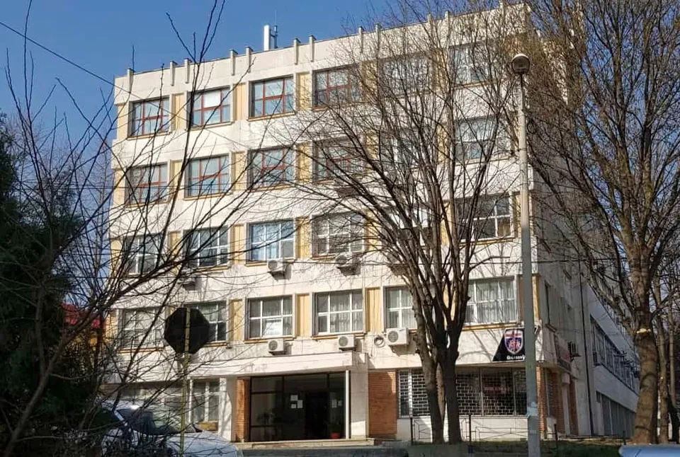 Un clădire cu patru etaje și ferestre multiple este parțial acoperită de ramurile unor copaci fără frunze sub un cer senin.