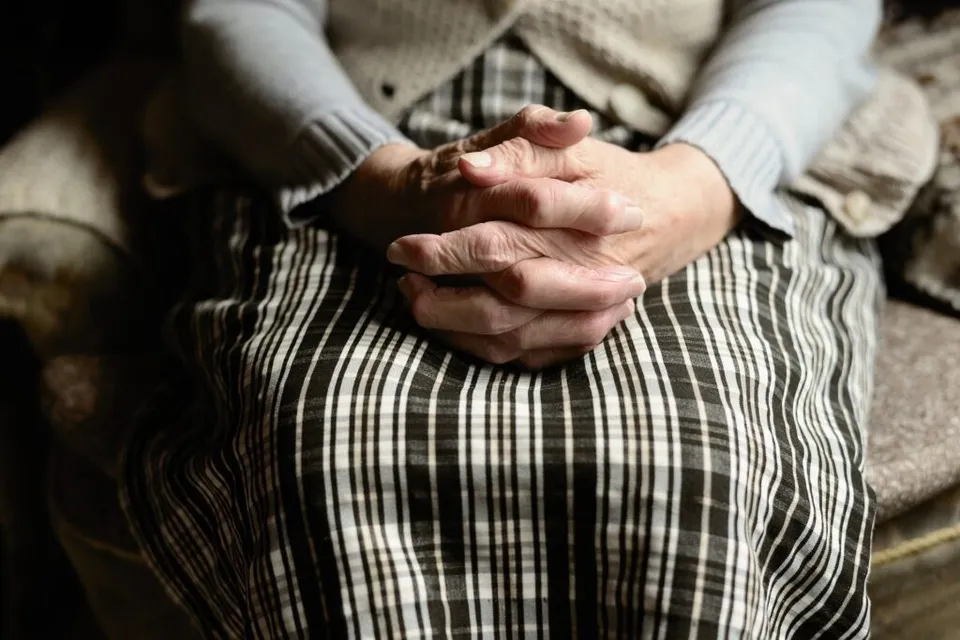 O persoană poartă un cardigan deschis peste o fustă cu model în alb și negru, având mâinile împreunate pe genunchi.