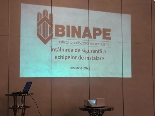 Proiecția unui logo și a unor text pe un perete deschis, lângă un laptop pe un suport și un proiector pe o masă.