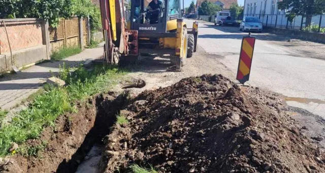 O excavatoare galbenă stă lângă un șanț deschis în pământ și un morman de toprak pe un drum asfaltat.