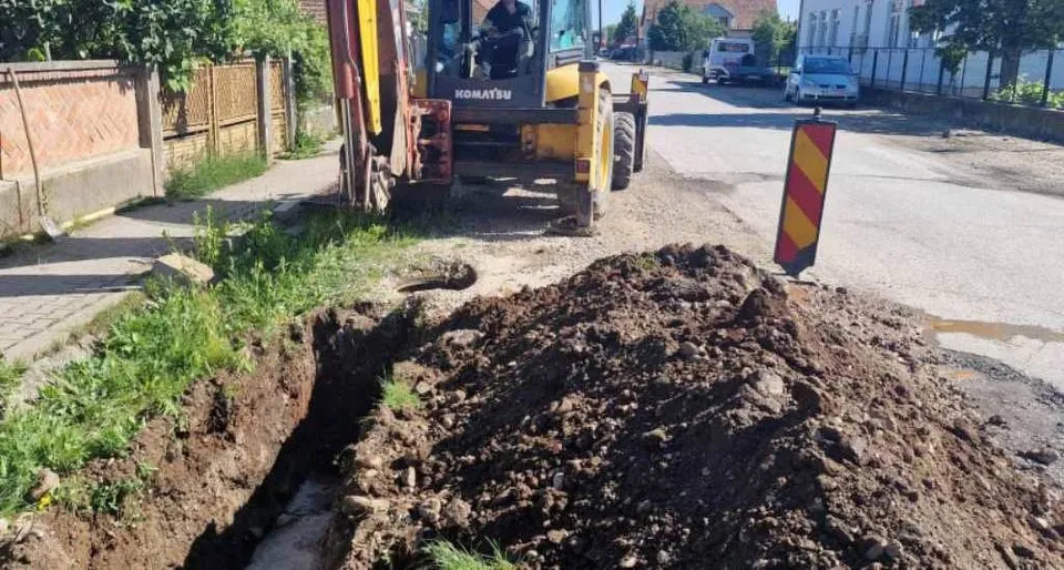 O excavatoare galbenă stă lângă un șanț deschis în pământ și un morman de toprak pe un drum asfaltat.