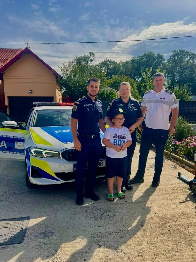 Trei polițiști în uniformă stau lângă un copil care poartă un tricou alb cu inscripții, în fața unei mașini de poliție.