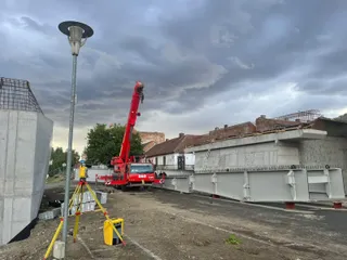Un utilaj de construcții roșu cu braț telescopic este poziționat lângă elemente de beton gri pe un șantier sub un cer înnorat.