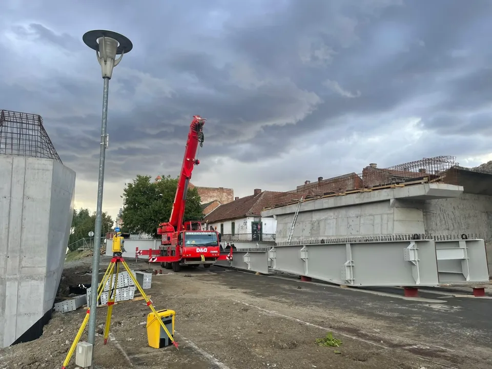 Un utilaj de construcții roșu cu braț telescopic este poziționat lângă elemente de beton gri pe un șantier sub un cer înnorat.