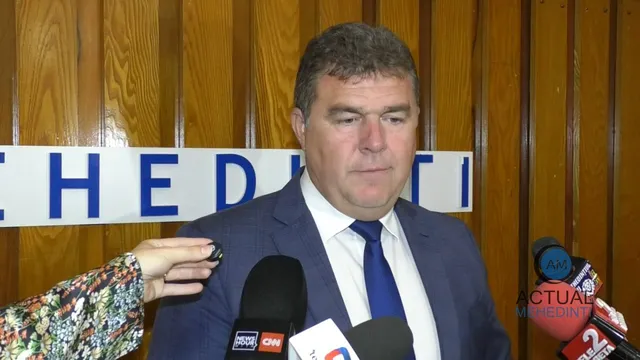 Un bărbat cu sacou și cravată albastră stă în fața mai multor microfoane de presă în fața unui panou cu litere albastre pe fundal alb.