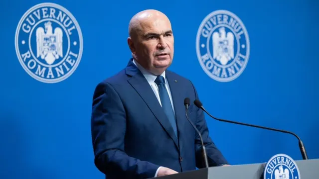 Un bărbat cu părul scurt și costum închis la culoare stă în fața unui fundal albastru cu două embleme circulare albe.