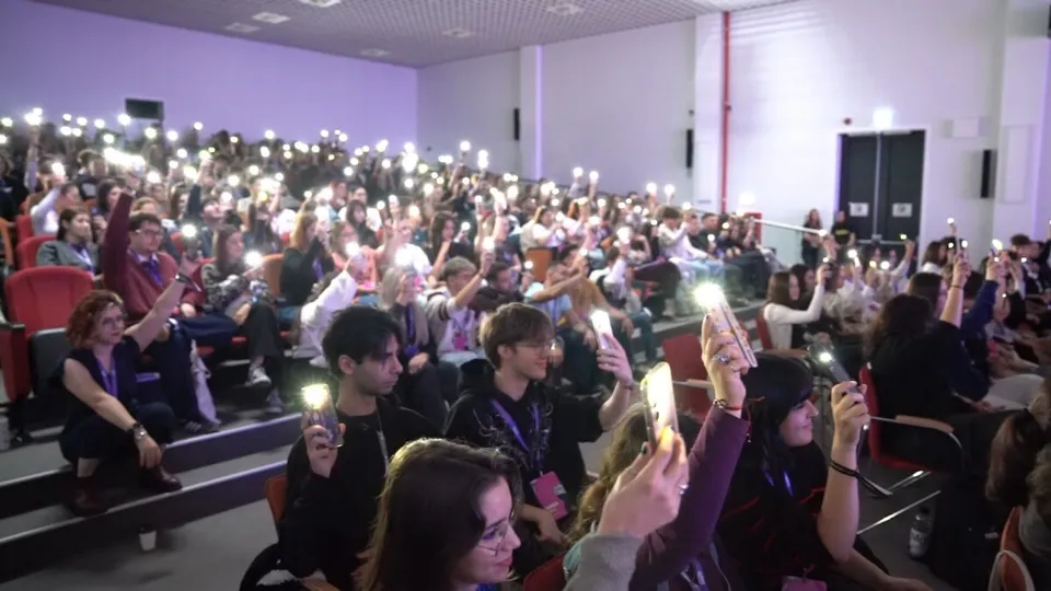 O mulțime de oameni stă așezată în scaune într-o sală de conferințe, ținând telefoane mobile cu lumina aprinsă în sus.