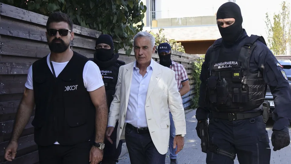 Un bărbat cu părul alb și sacou deschis merge între doi agenți în uniformă neagră și un bărbat cu vestă neagră și ochelari de soare.