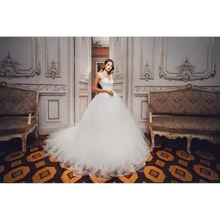O femeie poartă o rochie albă voluminoasă cu talie marcată, stând într-o încăpere cu panouri decorative și canapele de stil clasic pe un covor geometric.