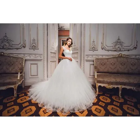 O femeie poartă o rochie albă voluminoasă cu talie marcată, stând într-o încăpere cu panouri decorative și canapele de stil clasic pe un covor geometric.