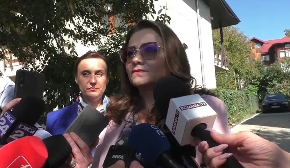 O femeie cu ochelari de soare și părul castaniu este înconjurată de microfoane de la mai multe stații de televiziune în exterior.