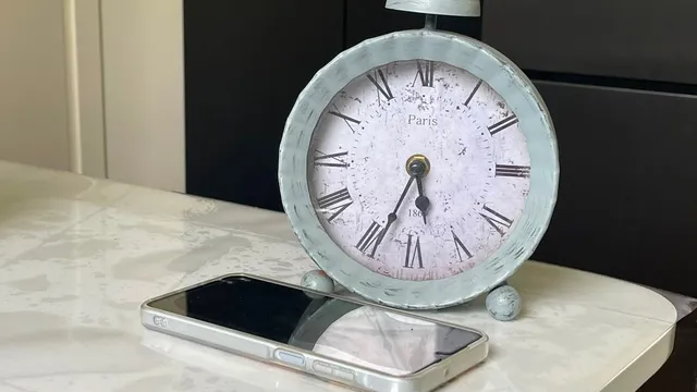 Un ceas analogic cu cifre romane stă pe o suprafață albă lângă un telefon mobil cu ecran negru.