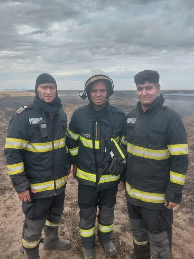 Trei bărbați îmbrăcați în uniforme de pompier negre cu benzi reflectorizante galbene stau în picioare pe un teren deschis sub un cer înnorat.