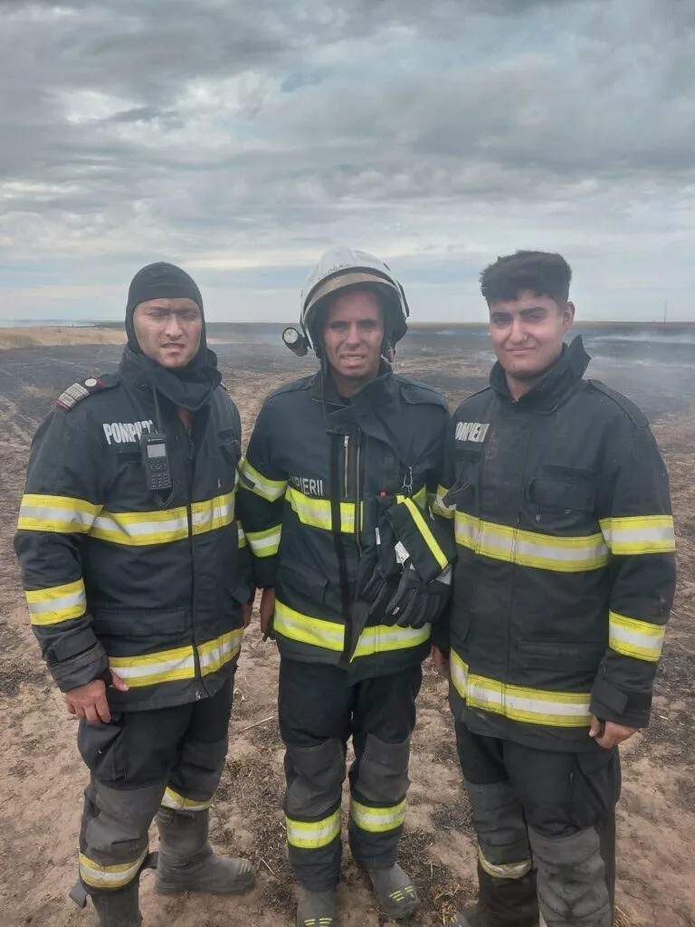Trei bărbați îmbrăcați în uniforme de pompier negre cu benzi reflectorizante galbene stau în picioare pe un teren deschis sub un cer înnorat.