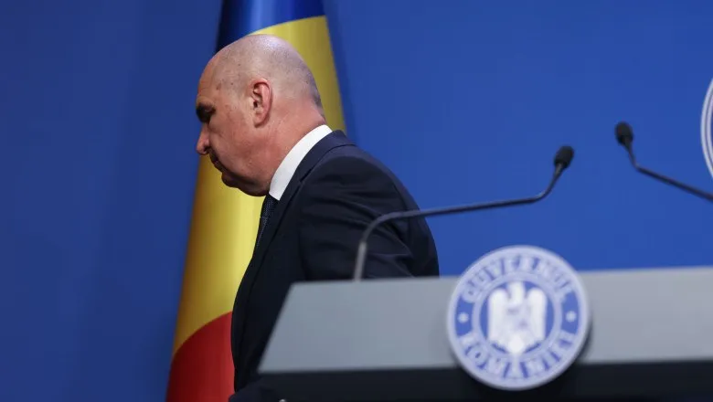 Un bărbat cu capul chel, purtând un costum închis la culoare și cămașă albă, stă în spatele unui podium cu un emblema circulară.