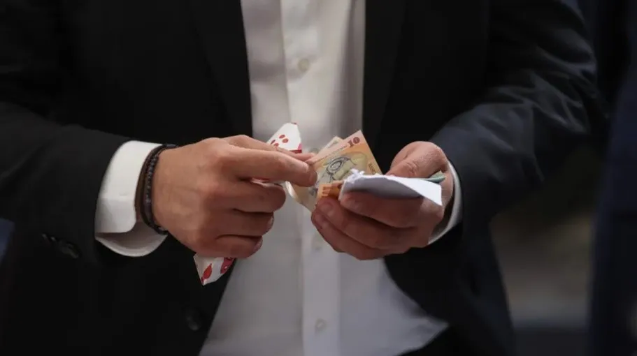 Un bărbat într-un costum negru și cămașă albă ține în mâini câteva bancnote de euro și un plic alb.
