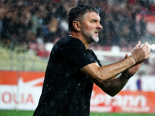 Un bărbat cu părul scurt și barbă căruntă, îmbrăcat în tricou negru, aplaudă sub ploaie pe un teren de fotbal.