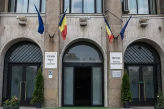 Fațada unei clădiri de piatră prezintă trei intrări cu arcuri, trei steaguri suspendate și două plăcuțe informative pe stâlpii de susținere.