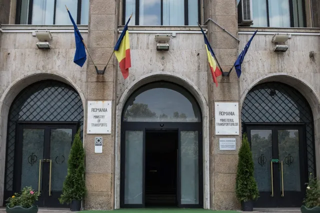 Fațada unei clădiri de piatră prezintă trei intrări cu arcuri, trei steaguri suspendate și două plăcuțe informative pe stâlpii de susținere.
