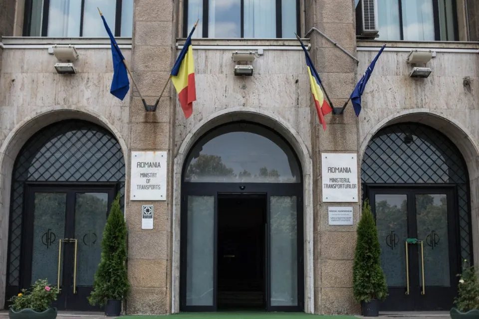 Fațada unei clădiri de piatră prezintă trei intrări cu arcuri, trei steaguri suspendate și două plăcuțe informative pe stâlpii de susținere.