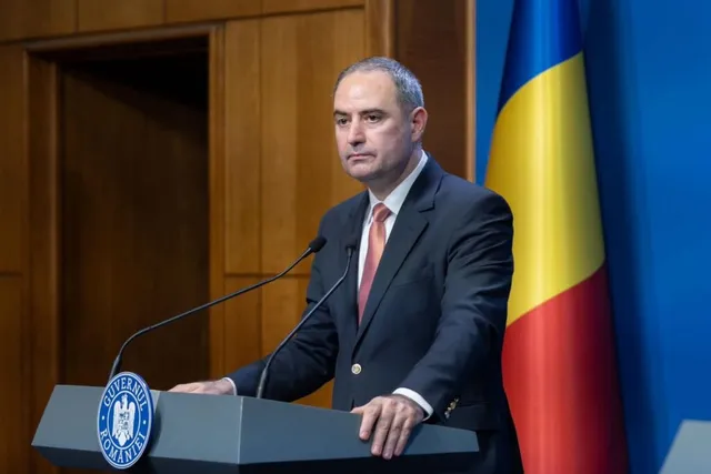 Un bărbat într-un costum închis la culoare și cravată roșie stă în spatele unui podium gri lângă un steag tricolor.