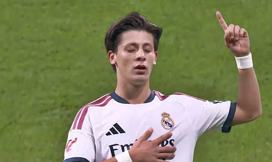Un jucător de Real Madrid în tricotul alb cu logo-ul echipei, cu brațul drept ridicat și arătând cu degetul, pe un fundal de iarbă verde.