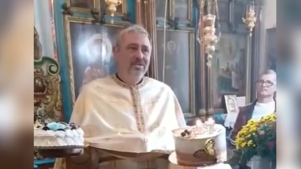 Un bărbat cu barbă, purtând o robă albă, stă în fața unor tablouri și a două prăjituri pe o masă.