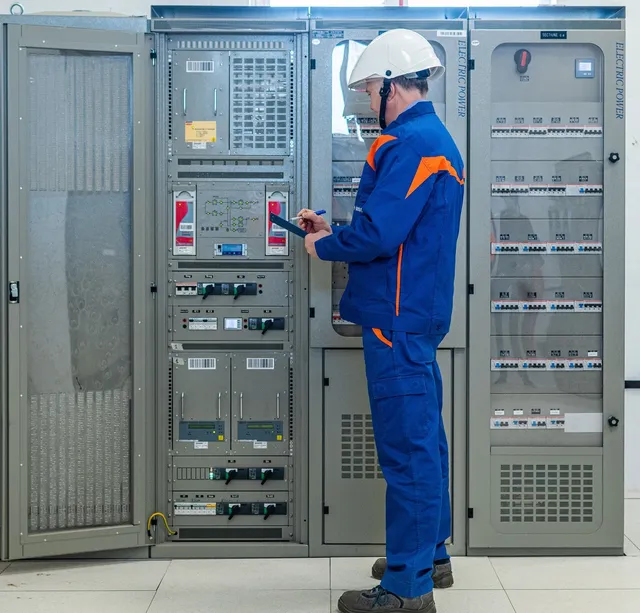 Un bărbat cu cască albă și costum de lucru albastru cu detalii portocalii scrie pe o foaie lângă panouri electrice gri deschise.
