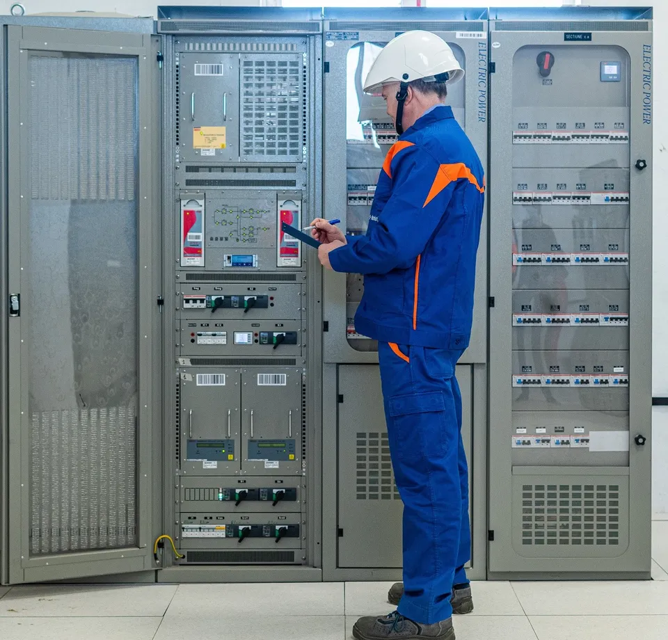 Un bărbat cu cască albă și costum de lucru albastru cu detalii portocalii scrie pe o foaie lângă panouri electrice gri deschise.